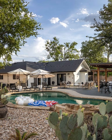 Modern home with pool, patio seating, umbrellas, and desert landscaping with cacti and mature trees
