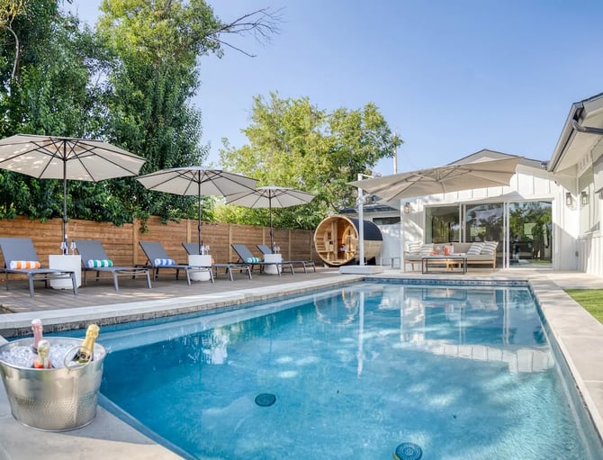 Backyard pool with lounge chairs, umbrellas, wooden fence, barrel sauna, and modern house with large windows